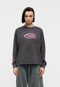 Νέος με κοντά σκοτεινά μαλλιά, φορώντας πλυμένο μαύρο φούτερ με ροζ κείμενο "THINK DIFFERENT" και φαρδιά γκρι παντελόνια, όρθιος.