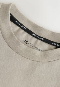 Bež bombažna majica z rahlo teksturiranim, okroglim izrezom. Ima črto etiketo z napisom "ALLSAINTS" in gladko površino tkanine.