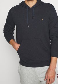 Haut à capuche gris foncé en mélange de coton, comportant une poche kangourou, une capuche réglable avec cordon et un petit logo doré sur la poitrine.