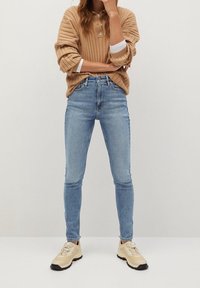 Jeans skinny de color azul claro combinados con un suéter naranja de canalé y zapatillas beige. El suéter tiene un cuello tipo polo y mangas anchas.
