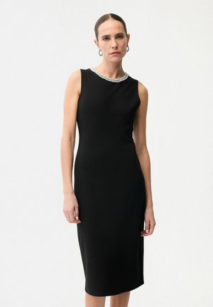 Vrouw die een mouwloze zwarte midijurk draagt met een kralenrandje bij de hals en hoepeloorbellen, staand tegen een effen witte achtergrond.