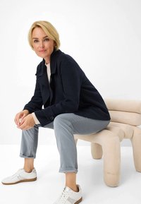 Marineblauwe wollen blend jas met een kraag, grijze broek met omgeslagen pijpen, witte sneakers, zittend op een beige sculpturale bank tegen een effen achtergrond.
