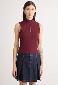 Haut sans manches bordeaux avec un col montant et une fermeture éclair à l'avant, associé à une jupe plissée en denim foncé avec des poches frontales et des détails de surpiqûres.