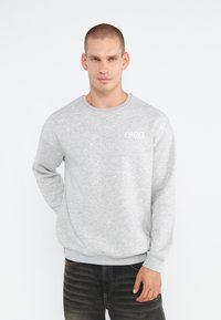 Grå sweatshirt med rund halslinning, lange ærmer og "PADEL" tekst på venstre bryst. Blød tekstur, parret med mørke jeans.
