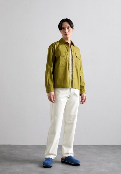 Olivno zelena lahka srajca z dvema sprednjima žepoma, v kombinaciji z belimi ravnimi kavbojkami in modrimi natikači. Nevtralna ozadje.