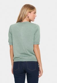 Kortärmad grön tröja i ett mjukt stickat material, med rund halsringning och ribbat avslut. Sedd bakifrån med mörkblå jeans.