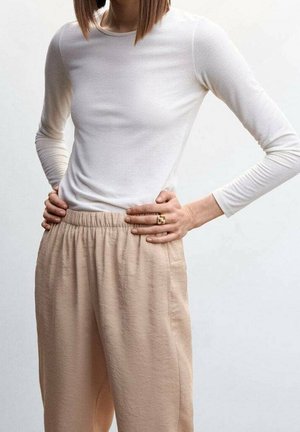 Personne portant une chemise blanche à manches longues et un pantalon beige à taille élastique, les mains posées sur les hanches, sur un fond clair uni.