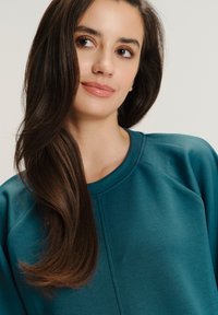 Tyrkysová mikina s kulatým výstřihem, měkkou texturou a raglánovými rukávy. Modelka má dlouhé vlasy, mírné vlny a přirozený make-up.