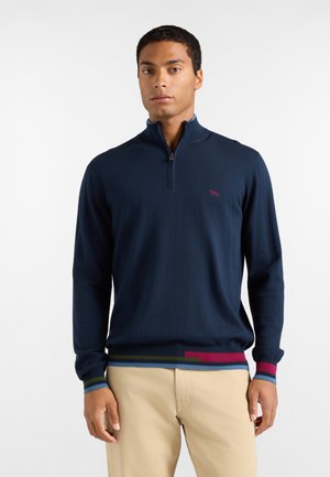 Uomo che indossa un maglione blu navy con zip a un quarto, con rifiniture colorate su polsini e orlo, abbinato a pantaloni beige, in piedi davanti a uno sfondo semplice.
