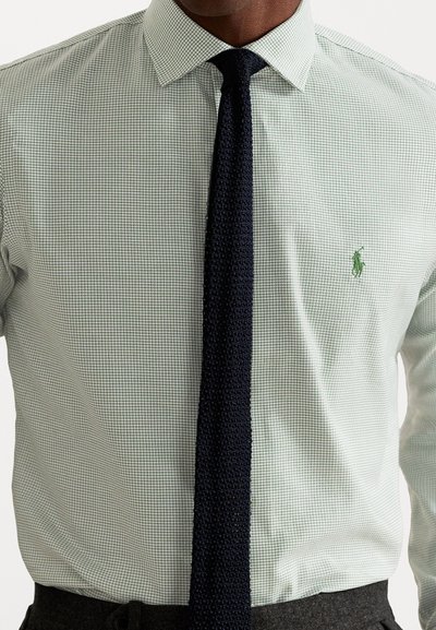 Bărbat îmbrăcat cu o cămașă de tip plaid în verde deschis, cu un logo brodat în verde și o cravată din tricot texturat în nuanță de navy, băgat în pantaloni închiși la culoare.