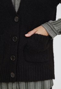 Svart stickad väst med ribbad nederkant, med fem knappar och två framfickor. Bärs över en grönrandig långärmad tröja.