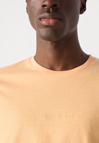 Tricou cu mânecă scurtă și guler rotund, din țesătură moale de piersică, cu text îmbosat "Arrivederci" pe partea din față, textură netedă și croială lejeră.