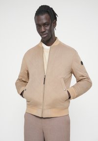 Μπουφάν τύπου bomber με καπιτονέ σχέδιο σε ανοιχτό μπεζ χρώμα, με ριμπ γιακά, κλείσιμο με φερμουάρ και πλαϊνές τσέπες. Διαθέτει λεπτομέρεια με μαύρο κουμπί στο αριστερό μανίκι.