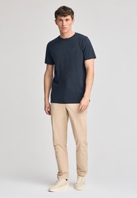 Czarna bawełniana koszulka z krótkim rękawem w kolorze granatowym, z małym kwadratowym logo na lewej piersi, połączona z beżowymi spodniami i jasnymi sneakersami.