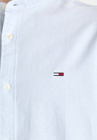 Ljusblå skjorta med knäppning, tillverkad av vävt tyg, med ståkrage och en liten broderad logotyp med röda, vita och blå accenter.