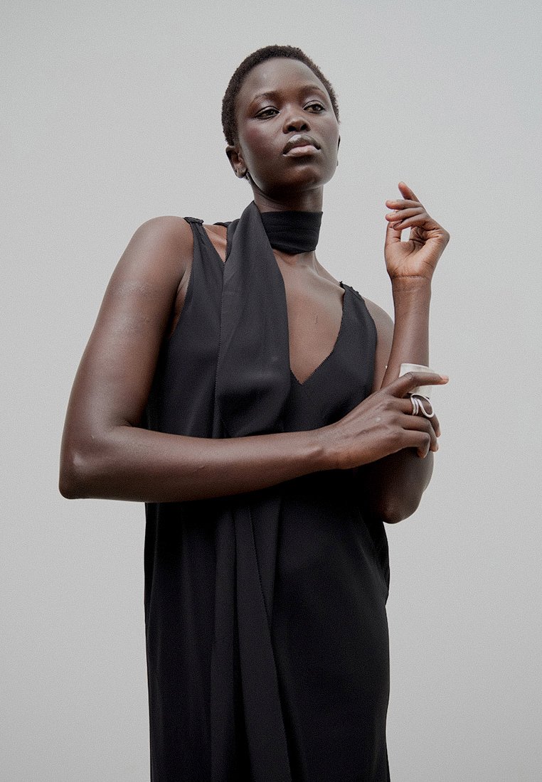 Czarna sukienka bez rękawów z głębokim dekoltem w szpic i drapowaną detalem przy szyi. Gładki materiał i dopasowany krój podkreślają sylwetkę. Prosty pierścionek na ręce.