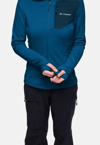 Giacca zip-up blu con una tasca sul petto di un blu scuro, realizzata in materiale morbido, con aperture per i pollici nelle maniche. Indossata con pantaloni neri.
