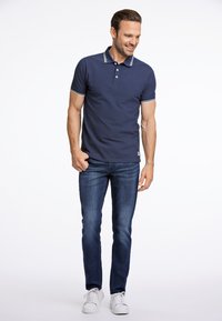 Mörkblå polo med vit kantning på kragen och ärmarna, matchad med mörka jeans och vita sneakers. Enkelt mönster och åtsittande form.