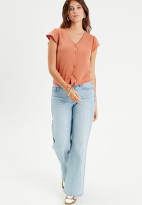 Blouse rouille à manches courtes avec un col en V et une fermeture à boutons, associée à un jean large bleu clair et des baskets beiges. Détails de design simples.