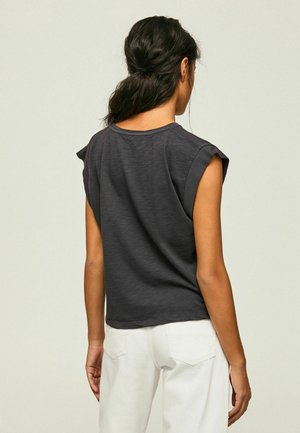 Kvinde med mørkt hår i en lav hestehale iført en mørkegrå ærmeløs top og hvide bukser, stående med ryggen til mod en ensfarvet baggrund.