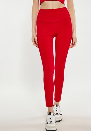 Røde leggings i slankt fit båret med hvide og sorte sportssko på en stående kvinde mod en ensfarvet lys baggrund.