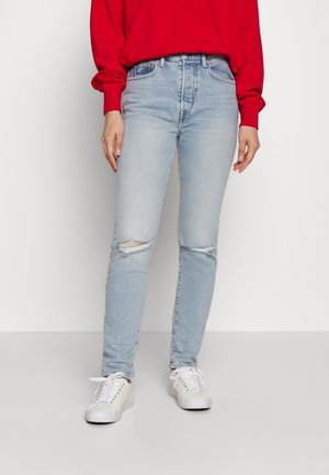 Osoba ubrana w jasnoniebieskie podarte obcisłe dżinsy, czerwoną bluzkę z długim rękawem i białe trampki, stojąca na jednolitym białym tle.