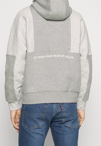 Person som bär en grå huvtröja med texten "KOM ÖVER DIN RÄDSLA FÖR HÖJDER" på ryggen, ihop med blå jeans.