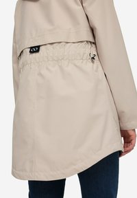 Ljusbeige regnjacka med samlad midja, elastisk dragsko, slät tyg och böjd fåll, som bärs över mörka jeans.