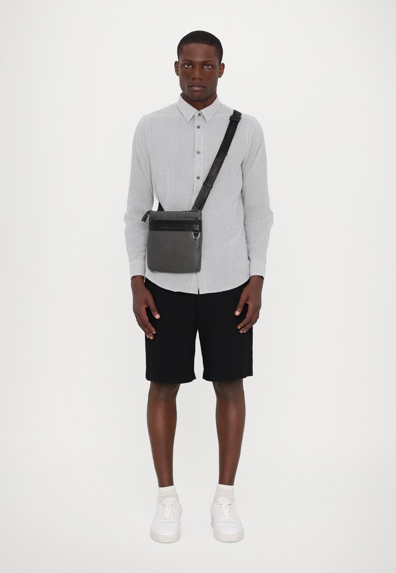 Borsa a tracolla in materiale nero, dotata di cerniera superiore, dettaglio del logo e tracolla regolabile. Il modello indossa una camicia a righe e pantaloni neri.