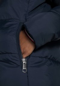 Námořnicky modrá prošívaná bunda s viditelným zipem, měkkou texturou a vnitřním lemováním manžety s červenými a bílými pruhy. Ruka částečně zasunutá v kapse.