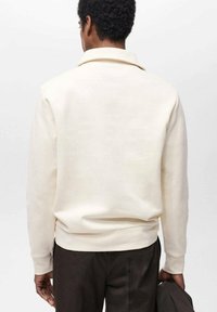 Felpa beige chiaro con colletto, tessuto testurizzato e polsini a costine. Presentata da dietro, abbinata a pantaloni marrone scuro.