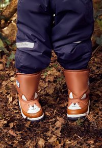 Stivali marroni, impermeabili, con un design di faccia di animale sorridente; accenti bianchi per gli occhi, forma strutturata; indossati sopra pantaloni blu scuro tra le foglie.