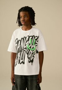 Weißes T-Shirt mit kurzen Ärmeln, das mit fettem schwarzen Graffiti-Text und einem grünen Smiley-Design versehen ist, kombiniert mit dunkelgrauen Cargo-Shorts.