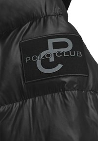 Svart vadderad jacka med matt finish, med en svart tygetikett sydd på axeln med texten "POLO CLUB" i grått.