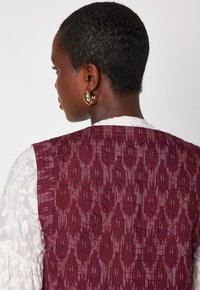 Donna con i capelli corti, che indossa orecchini a cerchio dorati, una blusa bianca ricamata e un gilet rosso con un motivo testurizzato, rivolta di spalle rispetto alla fotocamera.