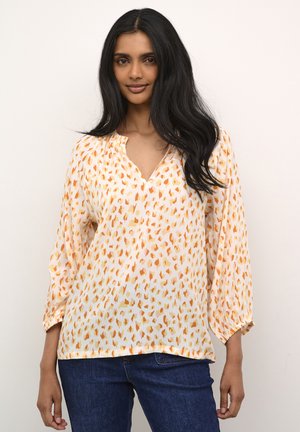 Vit blus med orange och gul abstrakt mönster, V-ringning, långa vid ärmar och samlat detaljer vid handleden. Kombinerad med blåjeans.