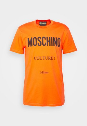 Henkilö, jolla on punainen Moschino Couture -t-paita, mustat jogger-housut ja mustat slip-on tennarit, joissa valkoiset pohjat ja Moschino-brändäys.