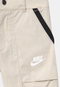 Beige kalhoty Nike s hladkým povrchem, se černým akcentem na boční kapse a výrazným bílým logem Nike v přední části.