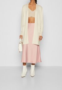 Lekki cardigan w kolorze kremowym założony na beżowy crop top, w zestawieniu z różową satynową spódnicą midi i białymi botkami za kostkę. Uzupełniony białą torebką.