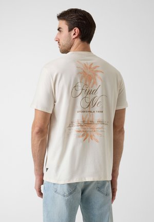 Homme portant un t-shirt écru avec un graphisme de palmier orange et le texte « Find Me Under a Palm Tree », associé à un jean bleu clair.
