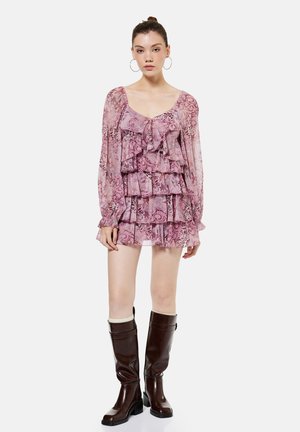 Mujer joven con vestido rosa de volantes con estampado y capas, de manga larga, y botas de cuero marrón hasta la rodilla, posando frente a un fondo liso.