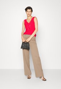 Top vermelho sem mangas com decote bordado, combinado com calças largas bejes. A modelo segura uma mala estruturada preta.