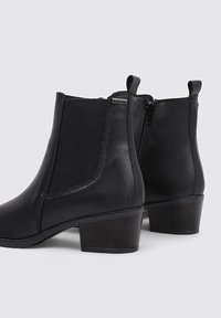 Black ankle boots made of leather with elastic side panels and a small block heel. Features a smooth texture and zipper closure.
