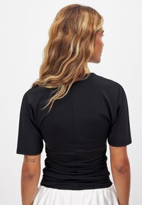 Juodas trumpų rankovių marškinėlis su prigludusia forma ir surinktu tekstūruotu nugaros dizainu. Lygus audinys, apvalus iškirpimas ir subtilūs siuvimo detalės.