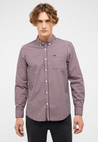 Młody mężczyzna w zapinanej czerwonej, granatowej i białej w kratę koszuli z długim rękawem, zestawionej z czarnymi spodniami, stojący na tle białego, jednolitego tła.