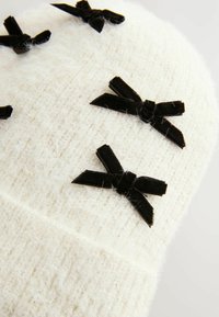 Bonnet tricoté blanc avec une texture duveteuse, ornée de plusieurs nœuds en velours noir comme embellissements.