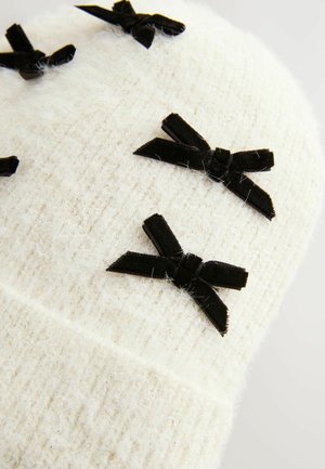 Bonnet tricoté blanc avec une texture duveteuse, ornée de plusieurs nœuds en velours noir comme embellissements.