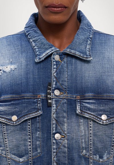 Denim jacket with a faded blue color, silver snap buttons, chest pockets, and a visible tag. Features distressed accents and prominent stitching.