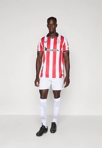 Fotbalový dres s červeno-bílými pruhy a černými akcenty, bílé šortky a bílé ponožky po kolena. Černé kopačky dokončují outfit.