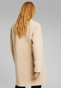 Beige ullblandad kappa med en mjuk, strukturerad yta. Har en avslappnad passform, långa ärmar och en längd till mitten av låret.